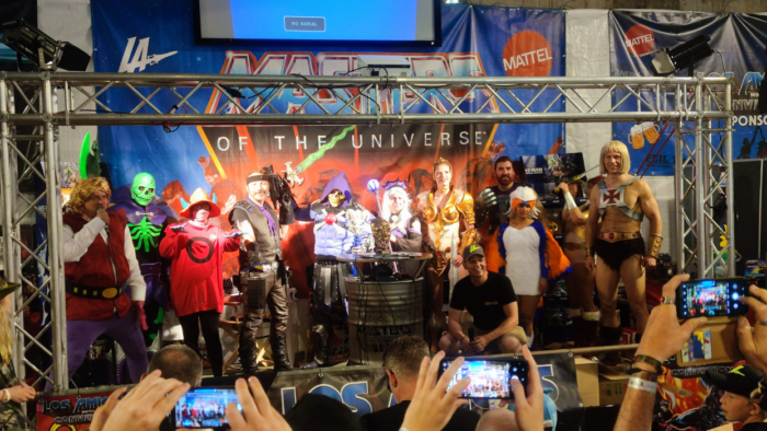 Eine Gruppe von Menschen in verschiedenen kostümierten Outfits posiert vor einem Bildhintergrund mit der Aufschrift 'Masters of the Universe' auf einer Veranstaltung.