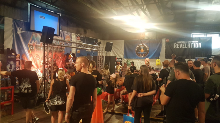 Ein großer Saal ist mit zahlreichen Menschen gefüllt, die sich vor einer Bühne versammelt haben. Die Atmosphäre ist entspannt und informell, viele Personen tragen schwarze Kleidung.