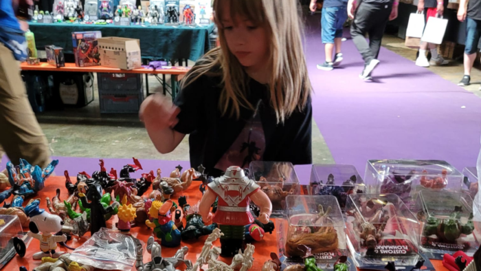 Ein Mädchen mit langem blonden Haar steht vor einem Tisch voller Spielzeugfiguren der Masters of the Universe-Reihe.