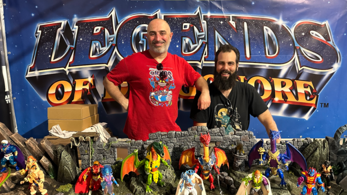 Zwei Männer stehen vor einer großen Bannerwand mit dem Aufdruck "LEGENDS OF Dragonore" in dunkelblauem Grundton und goldfarbenen Buchstaben, die sich hinter ihnen befindet. Sie selbst tragen T-Shirts mit diversen Designs und eine Sonnenbrille hängt an einem der Hemdkragen.