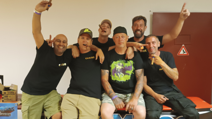 Sechs Männer sitzen auf einem roten Brett vor einer weißen Wand. Sie tragen schwarze T-Shirts mit gelben Schriftzügen. Einige von ihnen halten Zigarren in ihren Händen.