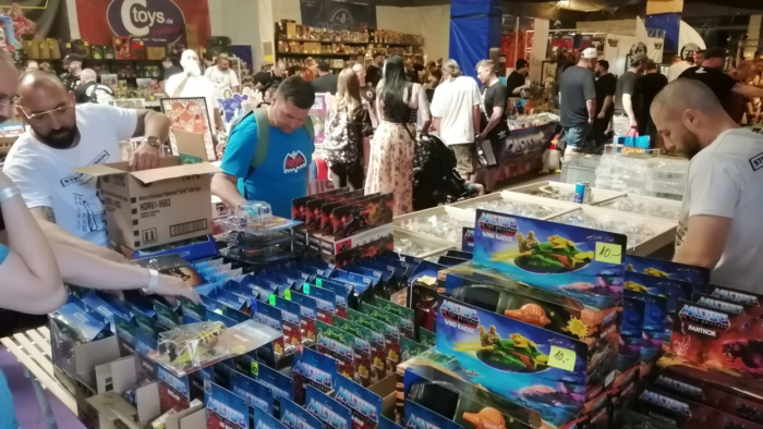 Eine Menge Menschen suchen sich auf einer großen Messe oder Ausstellung nach Spielen und Figuren aus der Welt von He-Man um. Viele Stände mit Schaufenster haben eine Vielzahl an verschiedenen Figuren, die in den Blister verpackt sind.